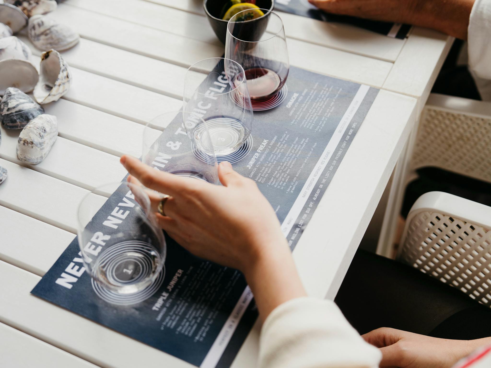 Hand holding tasting glass on gin flight mat with oyster shells on a white table.