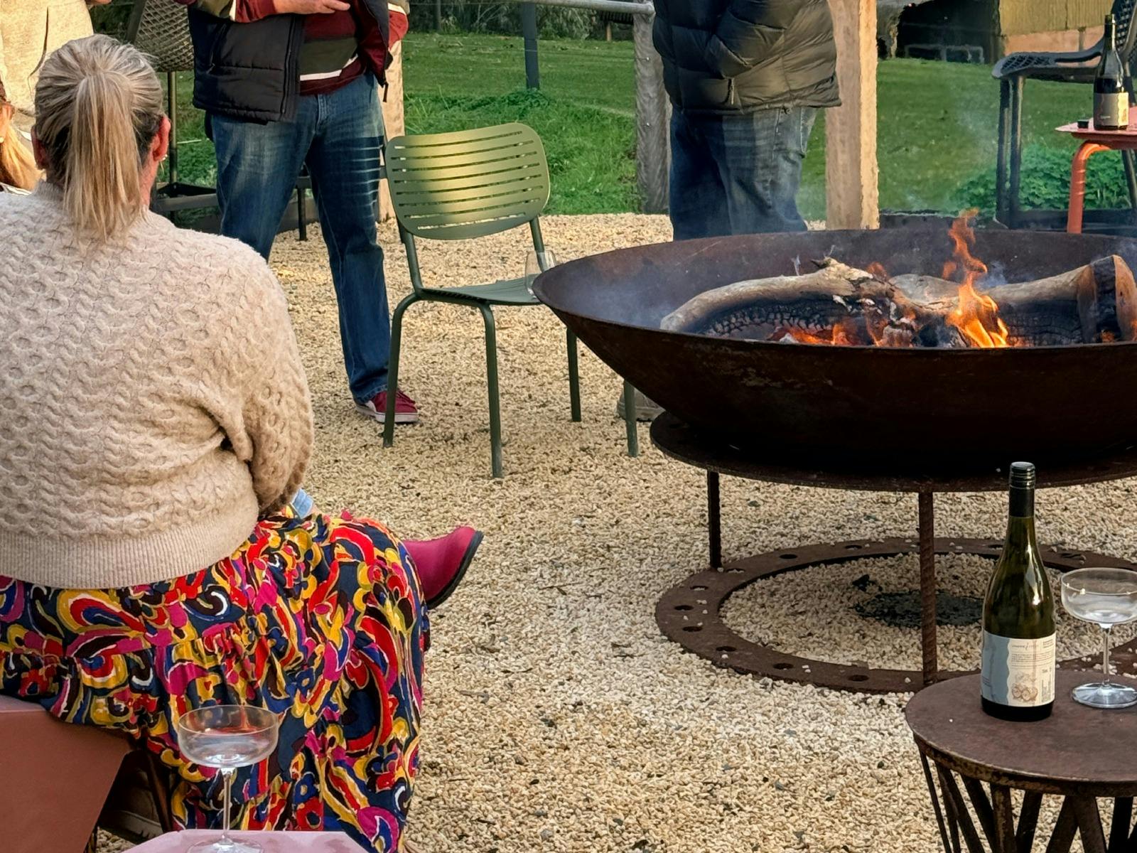 A bottle of wine in the foreground with people standing around a firepit