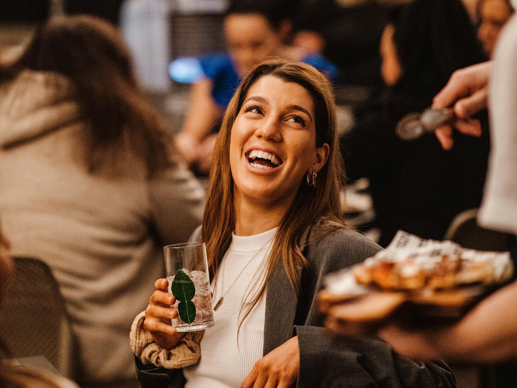 Woman in a gray suit laughs while holding a mint drink in a warm, lively, and cheerful setting.