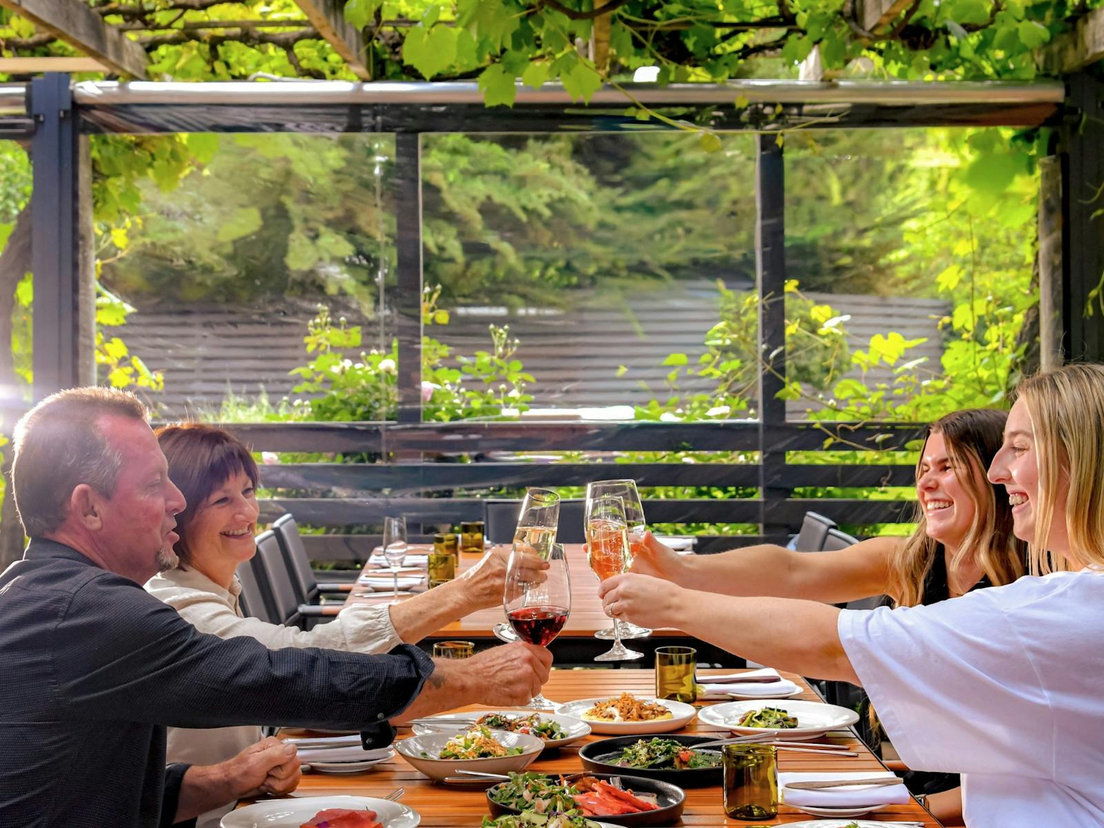 4 people clincking glasses over a table of food, beneath a vine covered arbor at The Currant Shed