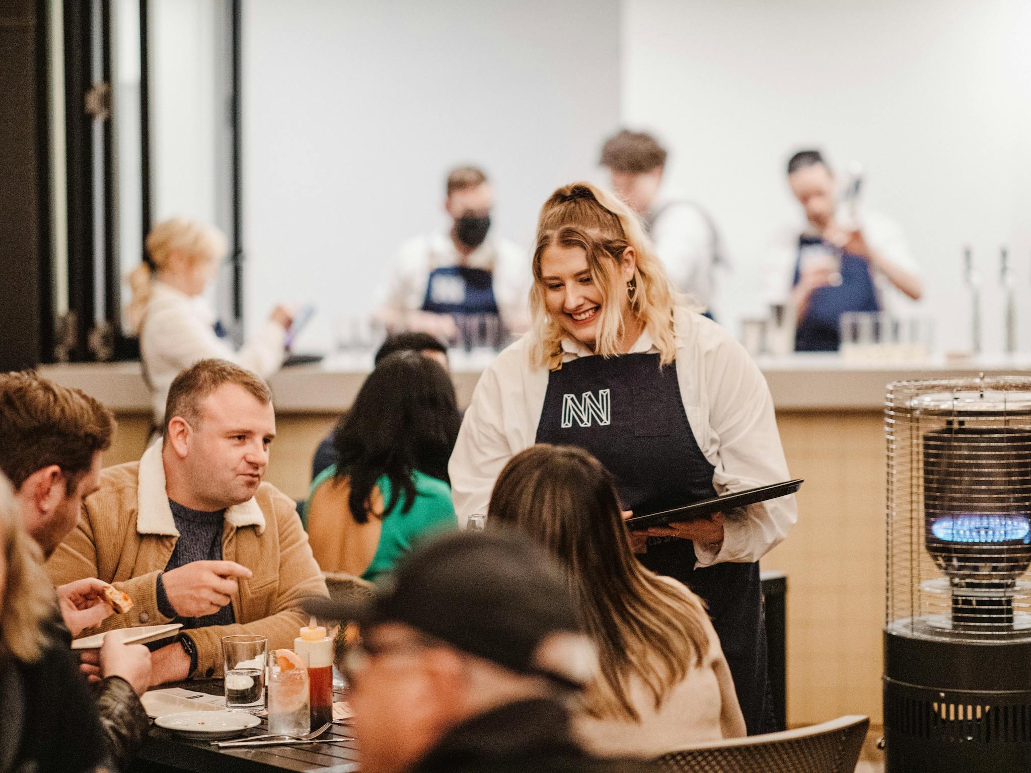 Cheerful waitress with menu approaches diners in a warm, lively distillery setting.