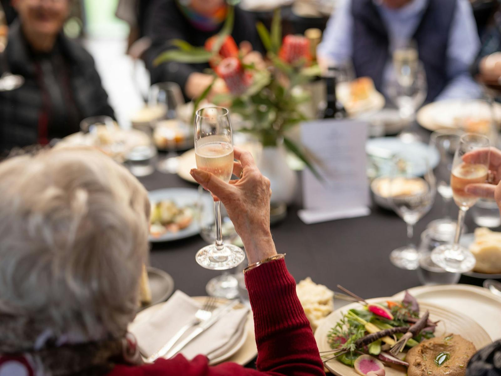 A table with food showing plates, vegetables, and people enjoying themselves with glasses of wine
