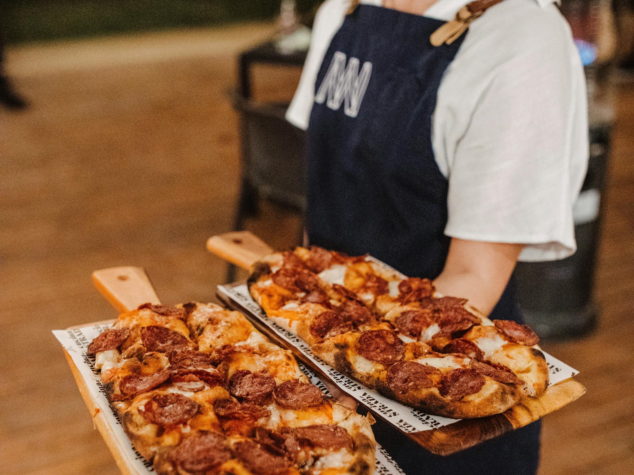 Gin expert in a dark blue apron carries two boards with freshly baked pizzas in a cozy setting
