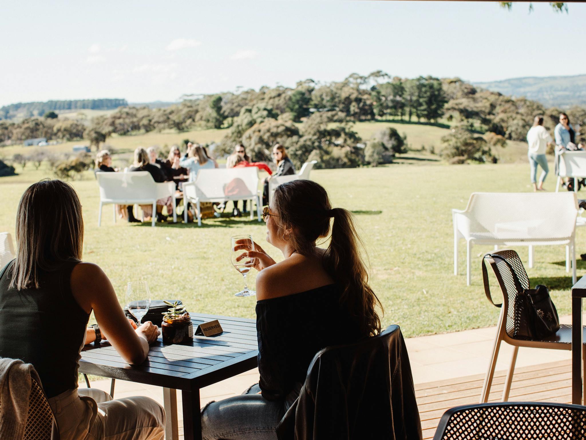 Two women sit at an outdoor table, enjoying drinks with a scenic view of rolling hills