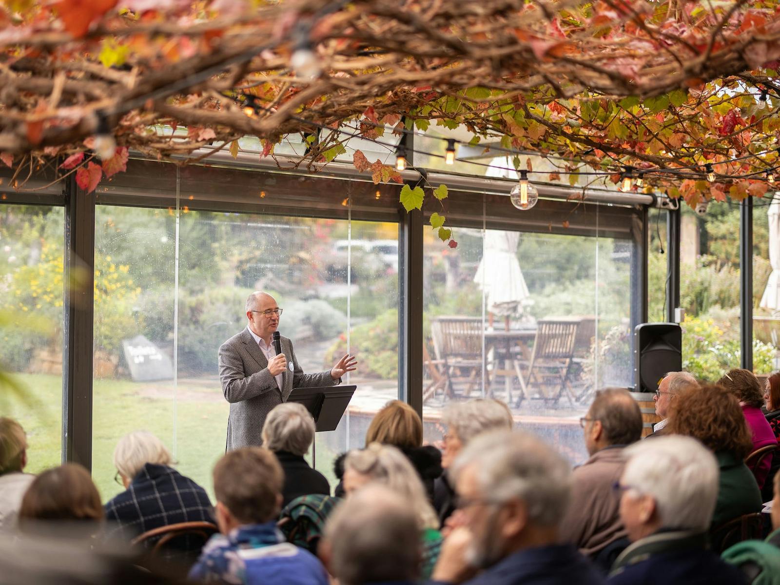 A man speaks in front of a crowd, standing under vine leaves in a courtyard
